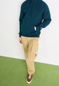 Felpa con cappuccio color verde petrolio con tasca a marsupio, abbinata a pantaloni cargo beige con multiple tasche. Scarpe da ginnastica marroni con suole bianche.