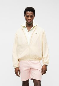 Νεαρός άνδρας φοράει κρεμ ζακέτα με φερμουάρ πάνω από ένα ανοιχτόχρωμο πουκάμισο με γιακά και παστέλ ροζ σορτς, στέκεται μπροστά σε ένα απλό λευκό φόντο.