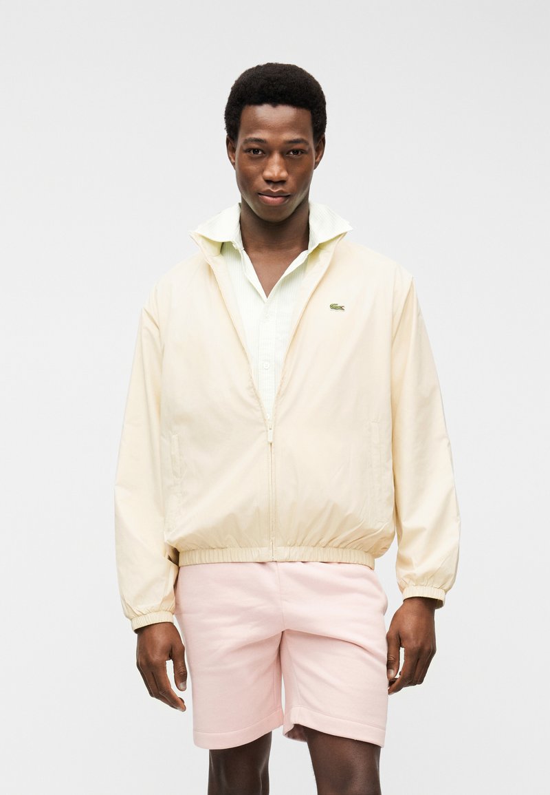 Νεαρός άνδρας φοράει κρεμ ζακέτα με φερμουάρ πάνω από ένα ανοιχτόχρωμο πουκάμισο με γιακά και παστέλ ροζ σορτς, στέκεται μπροστά σε ένα απλό λευκό φόντο.