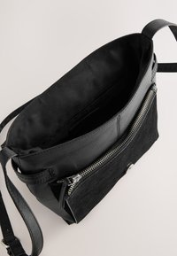 Sac bandoulière en cuir noir avec compartiment principal ouvert et poche avant zippée sur fond clair.