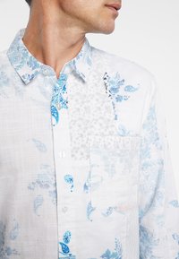 Camicia bianca con motivi paisley blu e accenti floreali, dotata di colletto button-down e una tasca applicata sul petto sinistro.