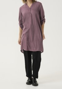 Tunique longue en tissu violet doux, dotée d'un col en V et de manches trois quarts. Assortie à un pantalon noir et des chaussures noires chunky.