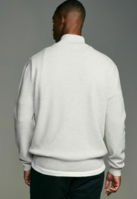 Grå fleecepolo, hög krage, ribbade mudd och nederkant, har en flerlagerslook med en vit skjorta under, mjuk textur.
