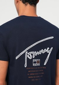 Sötétkék pamut póló, fehér és bronz nyomattal a háttal, rajta a "Tommy" logóval és városnevekkel rendezett elrendezésben.