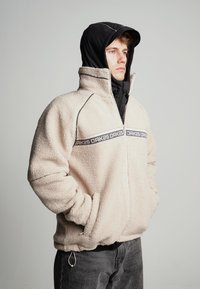 Jachetă de polar bej cu fermoar frontal, accente negre și o bandă cu logo. Dispune de glugă și ajustări elastice la margine și manșete.
