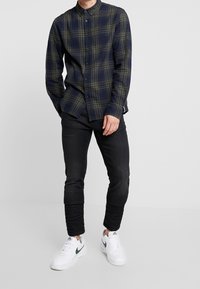 Mies, joka käyttää tummaa ruudullista napillista paitaa, mustia slim-fit farkkuja ja valkoisia Nike-tenniskenkiä, joissa on vihreä swoosh, seisoo yksiväristä taustaa vasten.