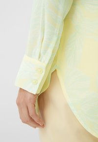 Blusa leggera gialla chiara in tessuto trasparente con sottili motivi a foglia blu, dotata di un polsino abbottonato e texture liscia. Ingrandimento della manica e della mano.