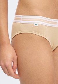 Μπεζ εσώρουχα με λευκή και μπεζ ριγέ μέση και ετικέτα με το λογότυπο της Lacoste, φορεμένα από ένα άτομο που φαίνεται από τη μέση μέχρι το μέσο του μηρού, σε στατική στάση.
