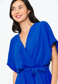 Modrá obalová šaty s kimono rukávmi a opaskom na páse. Obsahuje mäkkú, textúrovanú látku a hlboký výstrih do V.