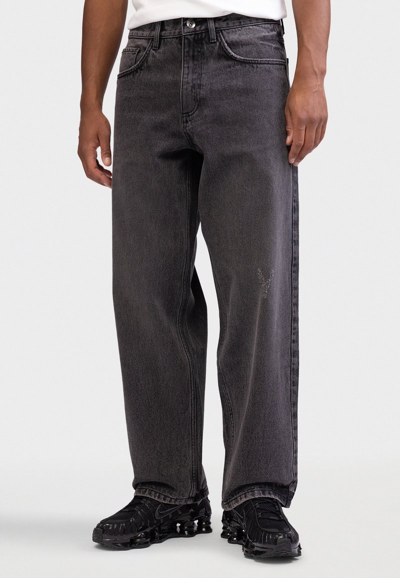 Homme portant un jean noir délavé et ample ainsi que des chaussures de sport noires, debout devant un fond clair uni.