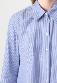 Blue striped blouse featuring vertical white stripes and small decorative silver studs, with a classic collar and button front.