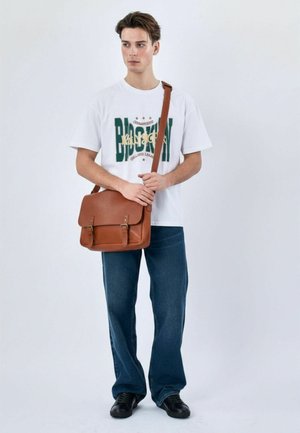 Junger Mann in weißem Brooklyn-Print T-Shirt, blauen Jeans, schwarzen Schuhen, der eine braune Lederschultertasche vor schlichtem Hintergrund hält.