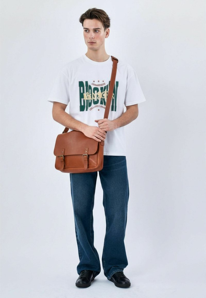 Jeune homme en T-shirt blanc imprimé Brooklyn, jean bleu, chaussures noires, tenant un sac à bandoulière en cuir marron sur un fond uni.