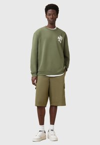 Felpa verde oliva con logo sul petto, abbinata a pantaloni corti coordinati e sneakers bianche. Tessuto in cotone misto, vestibilità rilassata.