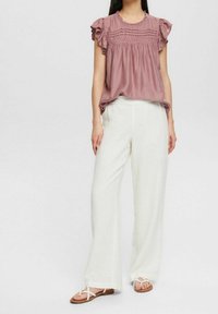 Lyserød, kortærmet bluse med flæseærmer og teksturerede detaljer kombineret med løse, hvide linnedbukser og flade sandaler.