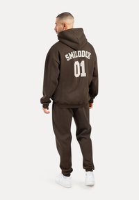 Brązowa bluza z kapturem z napisem "SMILODOX 01" na plecach, w połączeniu z pasującymi dresami i białymi sneakersami. Materiał wydaje się miękki i wygodny.