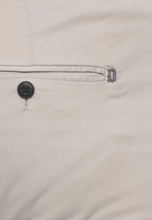 Tessuto beige con un piccolo bottone nero etichettato "Dondup" vicino a una cucitura orizzontale e un logo metallico a forma di "D" sul lato destro.