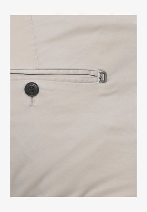 Tessuto beige con un piccolo bottone nero etichettato "Dondup" vicino a una cucitura orizzontale e un logo metallico a forma di "D" sul lato destro.