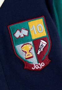 Naszywka haftowana z wzorem tarczy zawierającym zielone, żółte i bordowe sekcje; ikony błyskawicy, trofeum i gwizdka; tekst "10" oraz "Jojo".