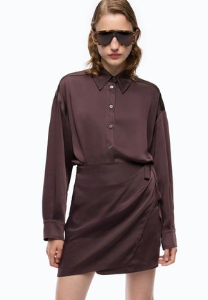 Mujer con blusa de manga larga color marrón oscuro abotonada y falda cruzada a juego, con grandes gafas de sol con montura carey, de pie sobre fondo blanco.