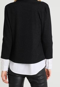 Svart, texturerad jacka med långa ärmar, ståkrage och kort längd, lager-på-lager över en vit skjorta och svarta läderbyxor.