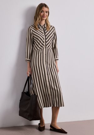 Kvinde iført en sort-hvid stribet midi kjole med knapper, der holder en stor mørkebrun lædertaske, står op ad en ensfarvet væg.