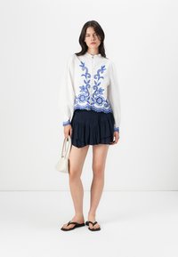 Blusa bianca con ricamo floreale blu, colletto rotondo, davanti con bottoni, abbinata a una mini gonna a balze blu navy e ciabatte nere. Borsa bianca.