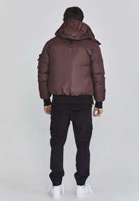 Braune Daunenjacke mit hohem Kragen und Kapuze, ausgestattet mit gesticktem Branding. Kombiniert mit schwarzen Cargo-Hosen und weißen Sneakers.