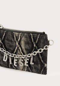 Μαύρο τσαντάκι από ντένιμ με καπιτονέ σχέδιο, ασημένια αλυσίδα ως λουρί και διακοσμητικά γράμματα που σχηματίζουν τη λέξη "DIESEL". Κλείσιμο με φερμουάρ στο πάνω μέρος.