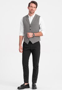 Chaleco de espiga gris sobre una camisa blanca, acompañado de pantalones negros ajustados y zapatos negros. Presenta un reloj en la muñeca izquierda.