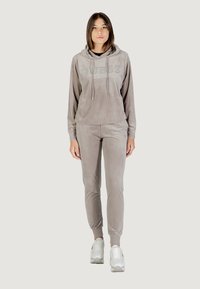 Chándal de terciopelo gris que incluye una sudadera con capucha con el logo "GUESS" y pantalones ajustados a juego con puños elásticos, combinados con zapatillas blancas.