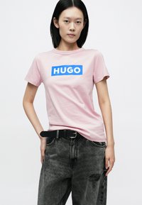 Lyserød kortærmet t-shirt med en blå rektangulær logo, der siger "HUGO" på forsiden. Modellen bærer sorte jeans og et sort bælte.