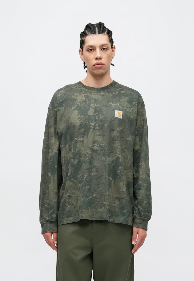 Young person with braided hair wears green camouflage long-sleeve shirt and olive pants, standing against a plain white background.