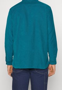 Camicia di velluto a coste color teal con maniche lunghe, polsini con bottone e taglio dritto. Tessuto morbido e dettaglio sullo yoke posteriore.