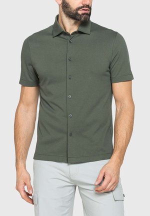 Uomo con barba che indossa una camicia verde oliva a maniche corte con bottoni e pantaloni grigio chiaro con tasche laterali, in piedi su uno sfondo semplice.