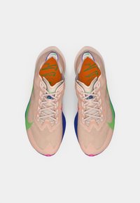 Buty sportowe z siateczkową cholewką w jasnym brzoskwiniowym kolorze, z zielonymi i niebieskimi akcentami na podeszwie oraz różowymi detalami na palcach.