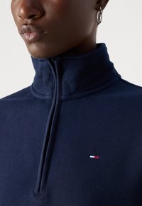 Navy half-zip sweater made of soft fabric with a high collar, featuring a small embroidered logo in red, white, and blue.