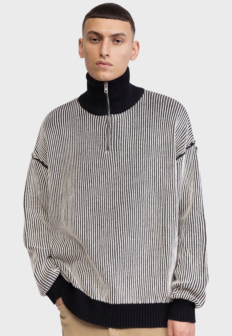 Pulover tricotat cu guler înalt cu fermoar, dungi verticale alb-negru și tiv și manșete cu textură ribbed.