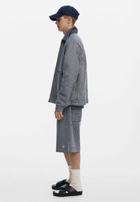 Csíkos sötétkék dzseki és rövidnadrág szett; laza szabás, textúrált anyag, oldalsó zsebek, sötétkék sapkával, bézs zoknikkal és fekete szandálokkal párosítva.