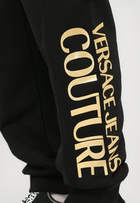 Must mustreev musta suurte kuldsete tähtedega "VERSACE JEANS COUTURE" vertikaalselt joondatud, koos musta kingaga, mis on osaliselt nähtav all.