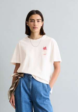 Jeune femme aux cheveux foncés portant un t-shirt blanc avec un motif graphique, un jean bleu taille haute, et une écharpe marron à motifs nouée à la taille, debout devant un mur gris.