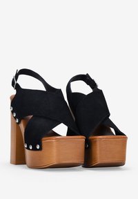 Zapatos de plataforma de ante negro con diseño de correas cruzadas y tacón de madera gruesa. Presenta tachuelas metálicas a lo largo del costado. Correa ajustable en el tobillo.