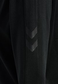Pantalones deportivos negros con una tela texturizada. Presenta dos acentos en forma de chevron negros en los laterales como detalle de diseño.