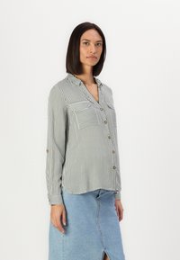 Camicia a righe verde e bianca con due tasche sul petto e bottoni dorati, abbinata a una gonna in denim azzurro chiaro.