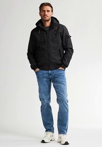 Zwart jack met ritssluiting en capuchon, voorzien van een ritssluiting aan de voorkant, zijzakken en een accentzak op de mouw. Draagt blauwe spijkerbroek en sneakers.