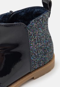 Lśniący, granatowy lakierowany skórzany botek z błyszcząca, wielokolorową teksturowaną boczną częścią i miękkim, ciemnym wyściółką wewnętrzną, z płaską, brązową podeszwą.