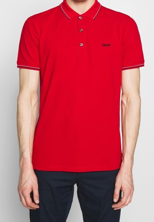 Polo rouge à manches courtes, patte de boutonnage à trois boutons, logo noir sur la poitrine, col et poignets de manches à rayures contrastées.