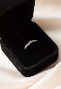 Bague en argent avec une pierre centrale transparente, présentant des bandes courbes ornées de petites pierres d'accent, exposée dans une boîte en velours noir sur un tissu doux.