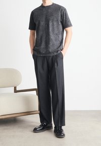 Svartprickig t-shirt med rund halsringning, ihop med svarta veckade byxor och glänsande svarta skor, står bredvid en beige stol.