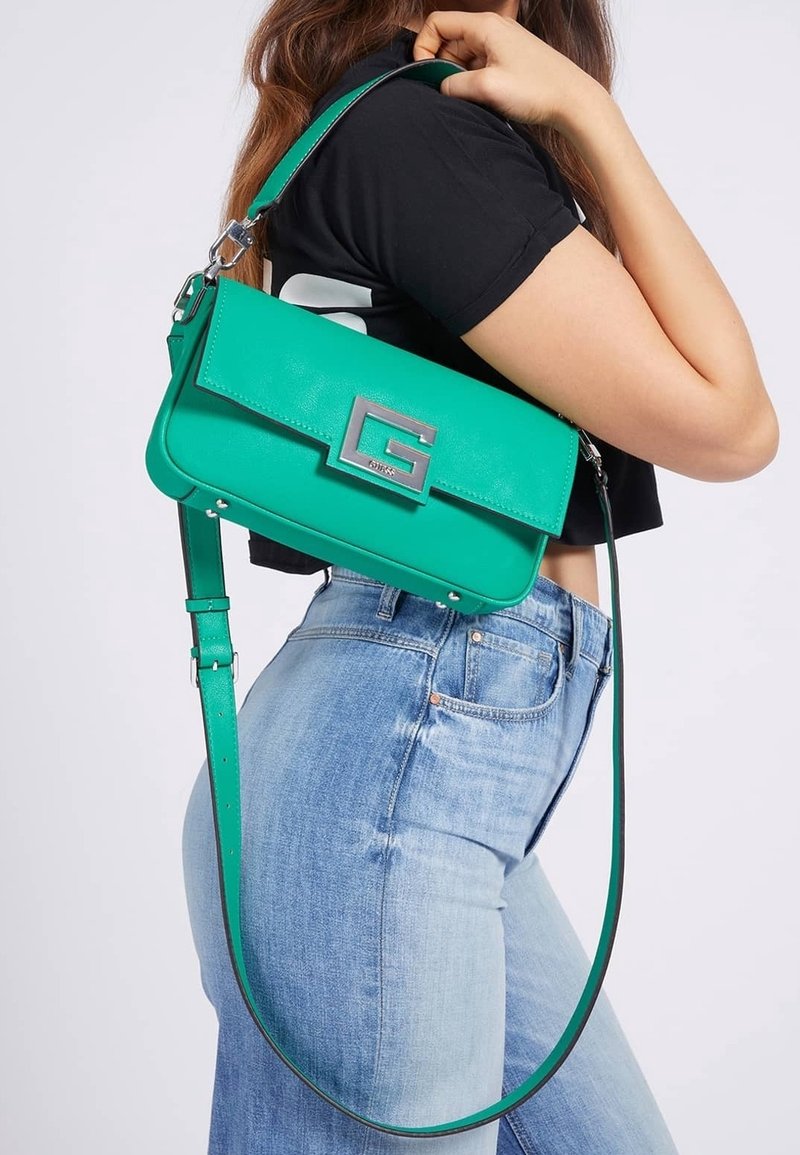 Personne portant un jean bleu clair et un haut noir, portant un sac à main vert avec un logo argenté "G" sur l'épaule.
